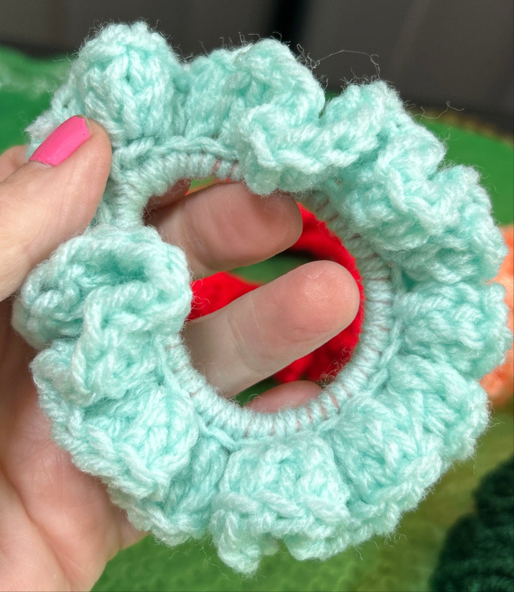 Hand holding a light blue knitted scrunchie with a red center against a green background.