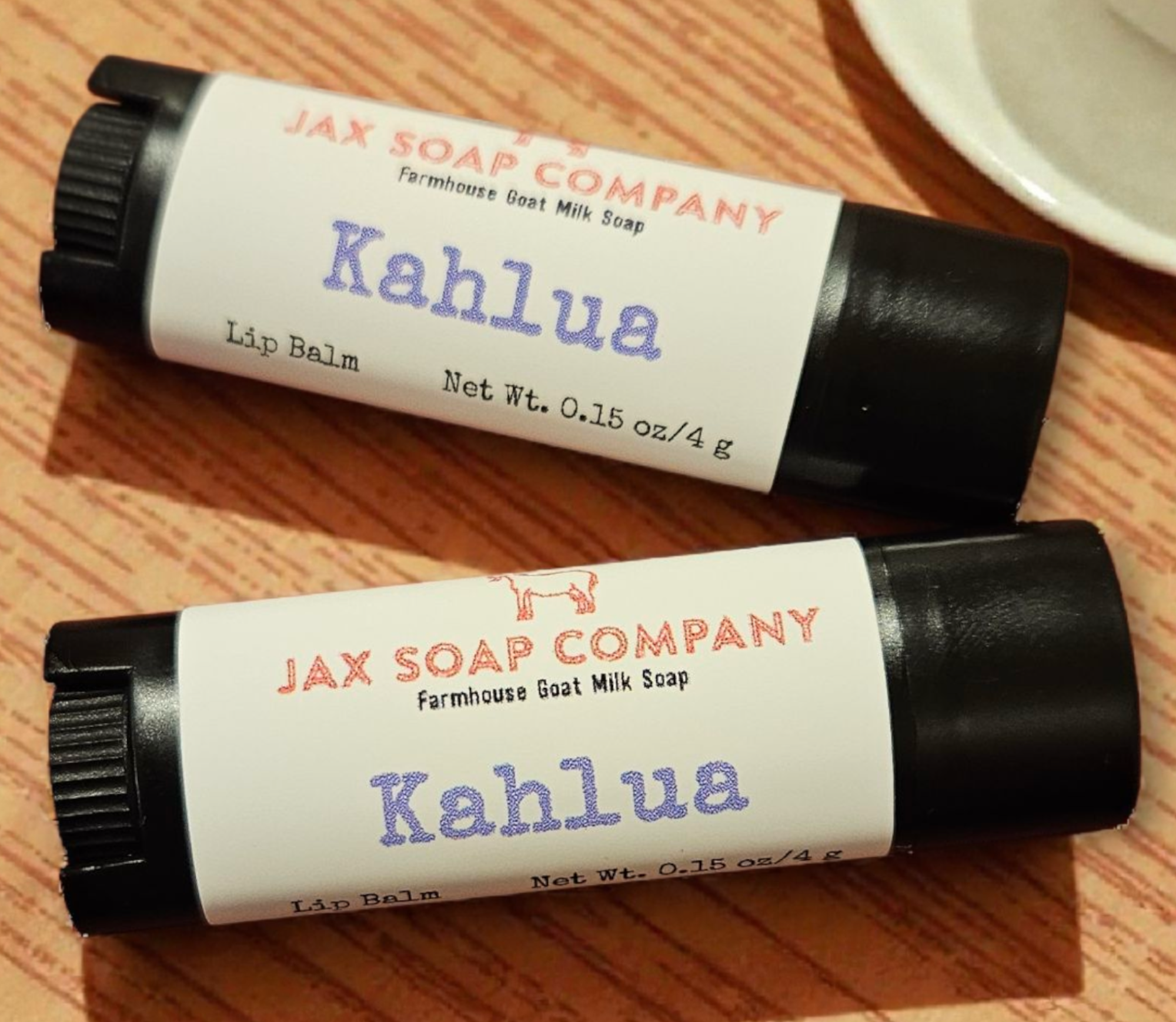 Two Jax Soap Company Kahlua lip balms on a wooden surface with a cup of coffee in the background.