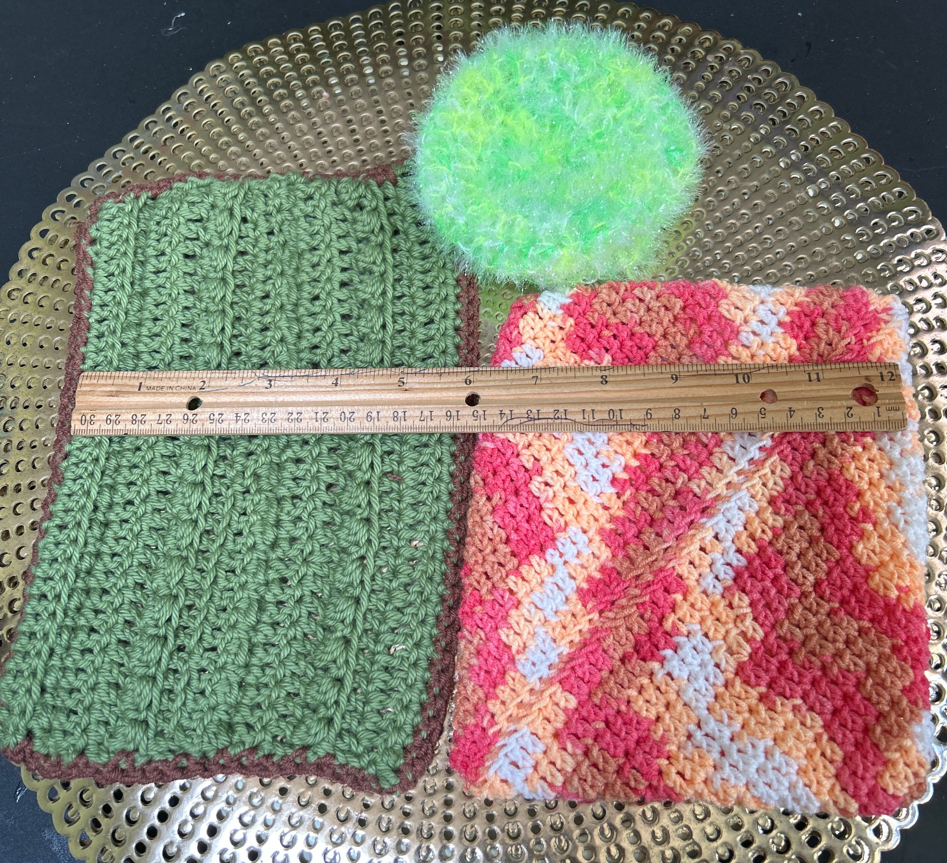 Two crocheted squares and a green pom-pom on a textured surface with a ruler for scale.
