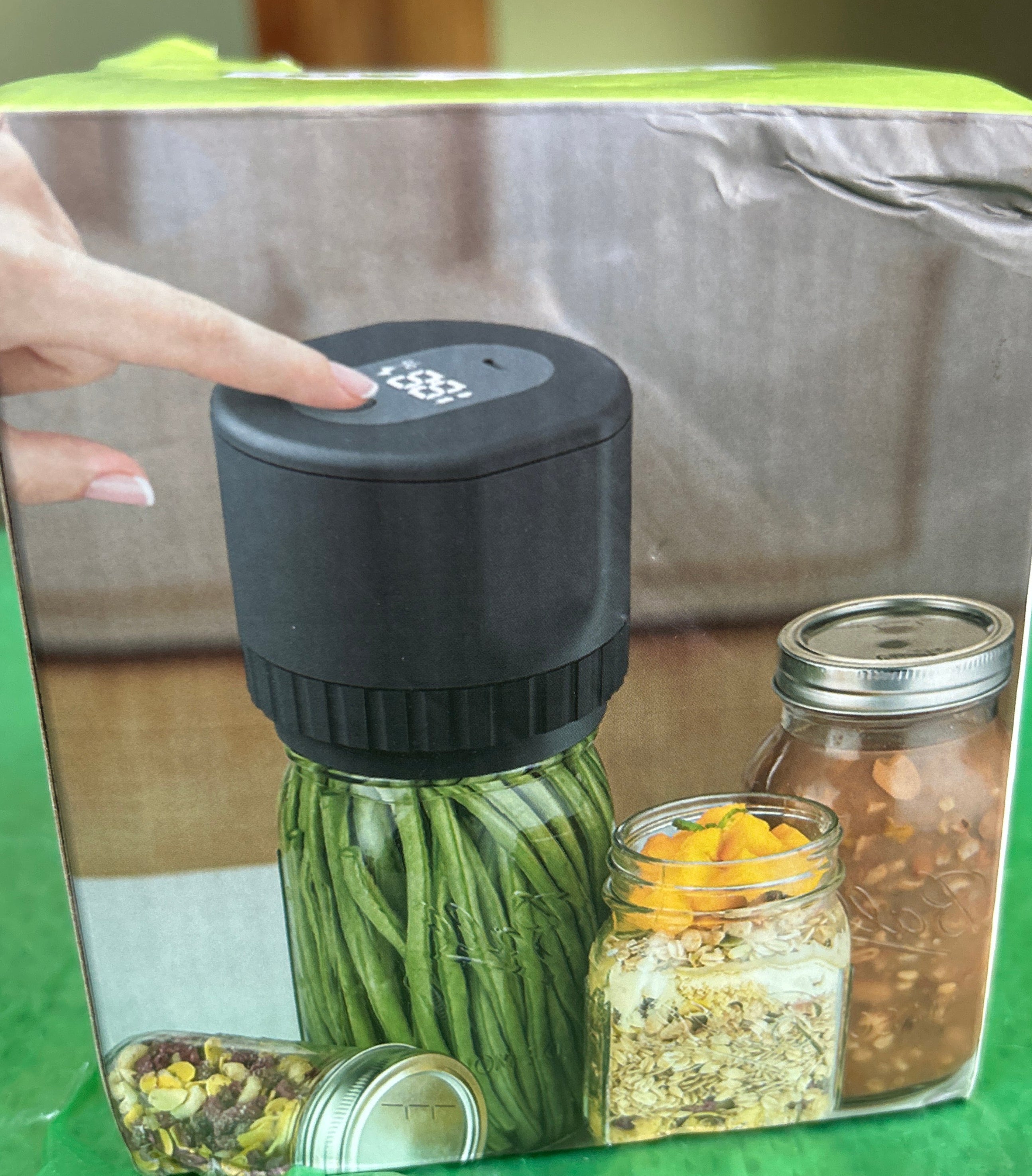 Product packaging with a hand pointing to a black cylindrical item, on a green surface.