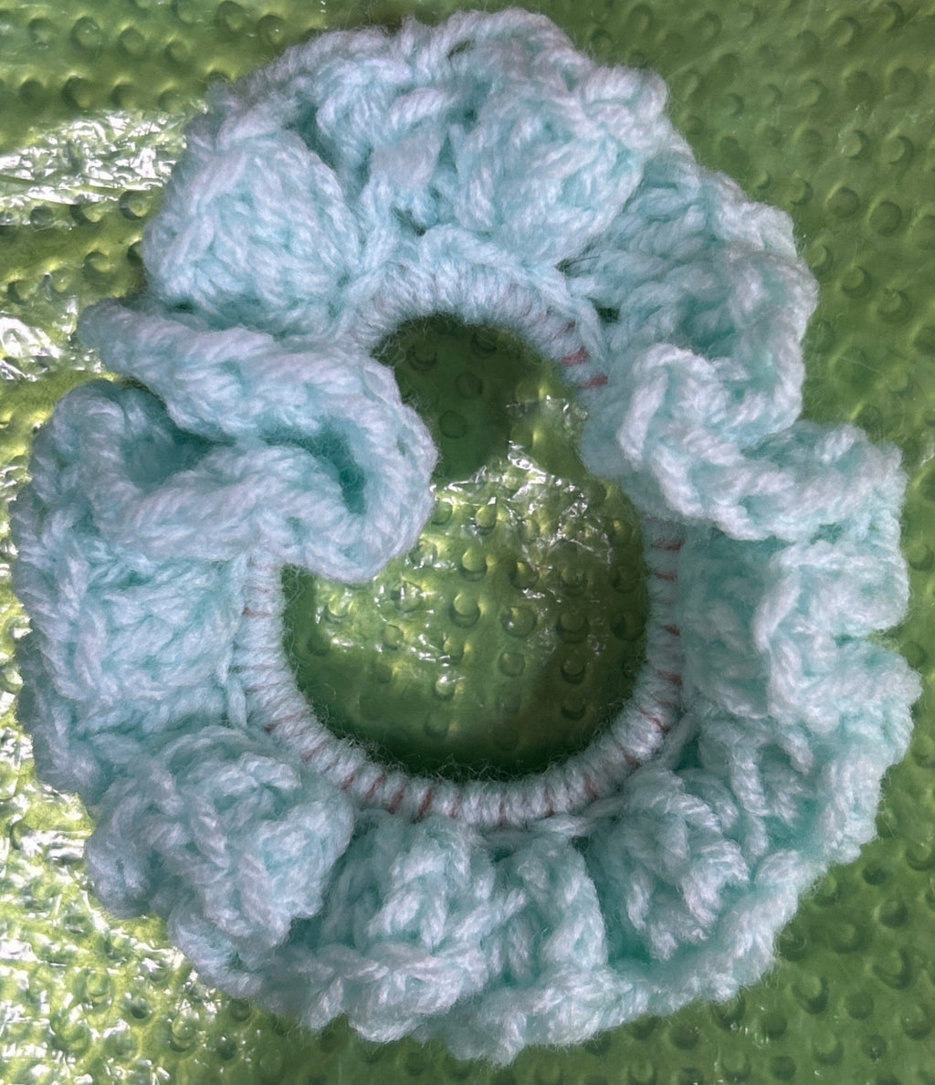 Blue crocheted scrunchie on a textured green surface with pink and red items partially visible.