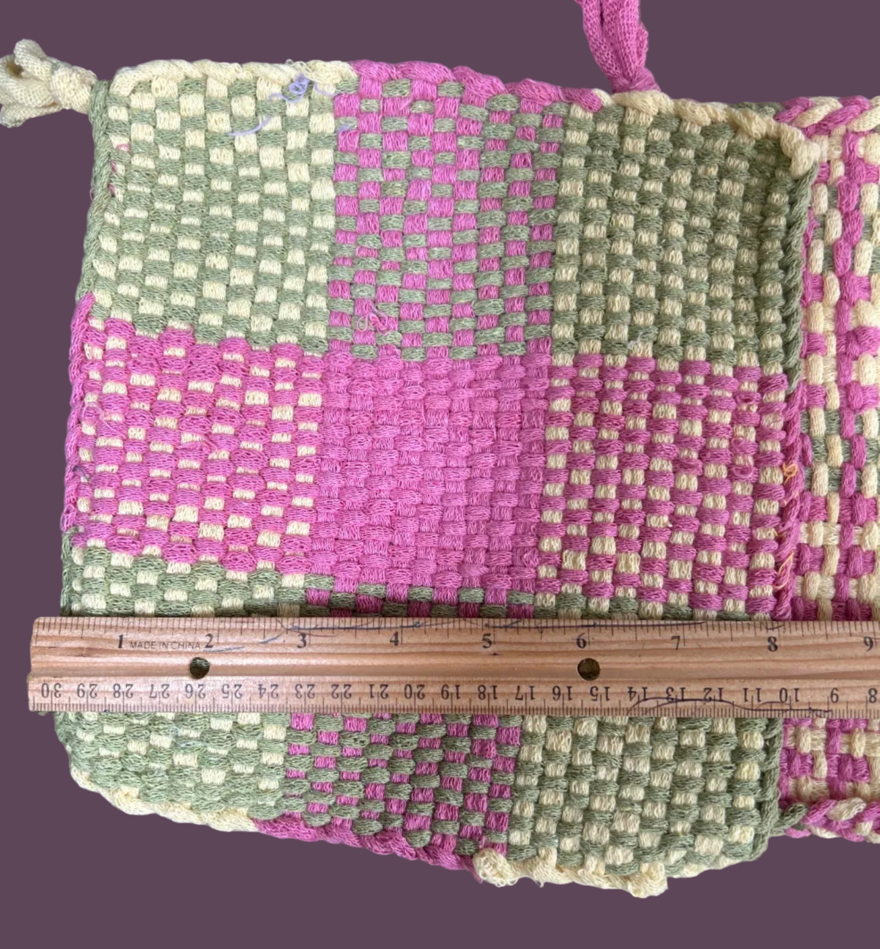 Handwoven pouch with pink and green pattern on a textured surface, with a ruler for scale.