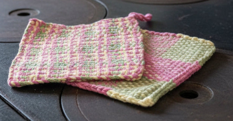 Hand-woven pink and green hot water bottle cover on a metal surface.