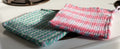 Two woven potholders, one pink and blue, one green and white, on a metallic surface.