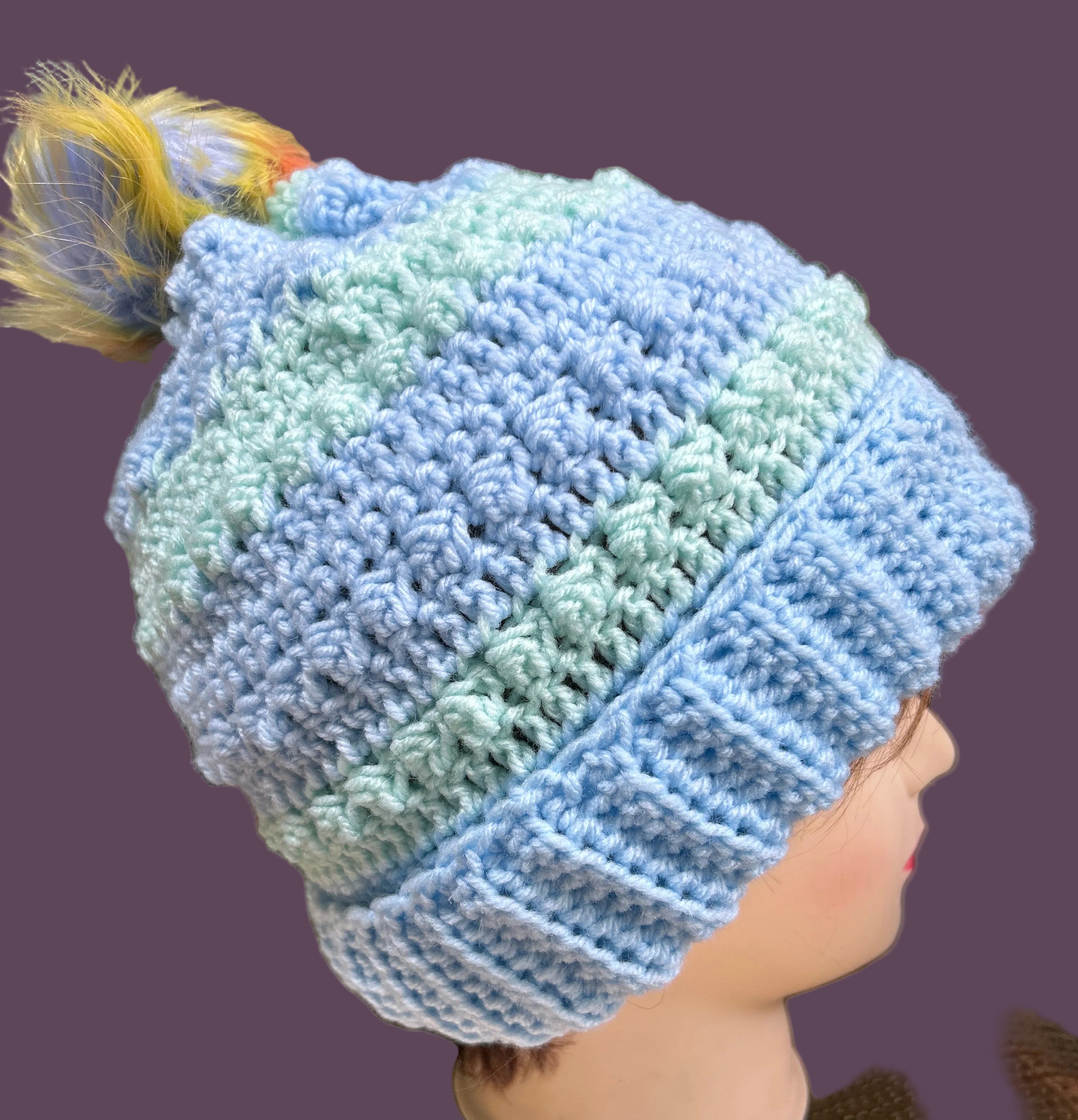 Close-up of a hand wearing a blue and green knitted beanie with a blurred background.