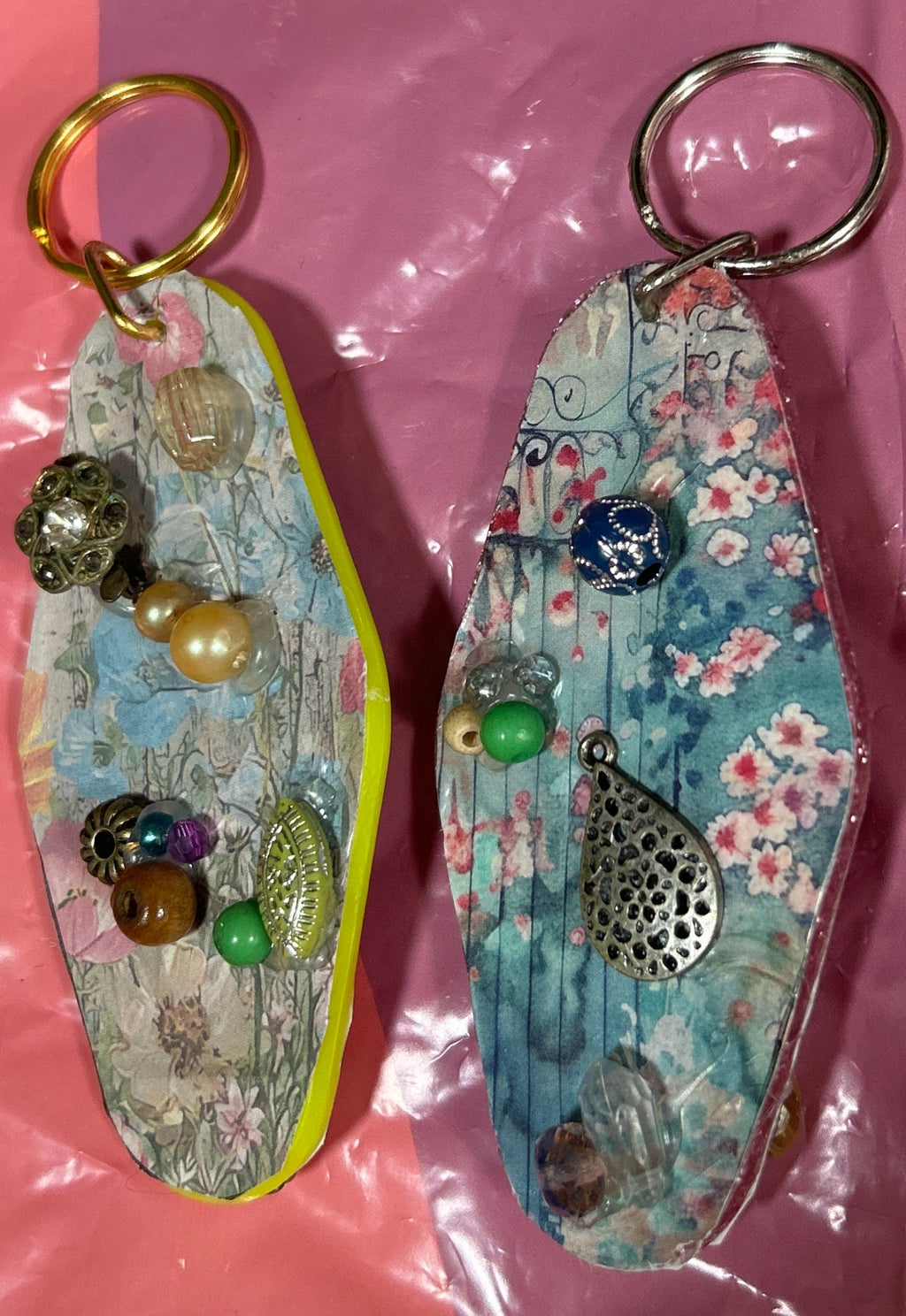 Two decorative keychains on a pink surface with a clear plastic bag in the background.