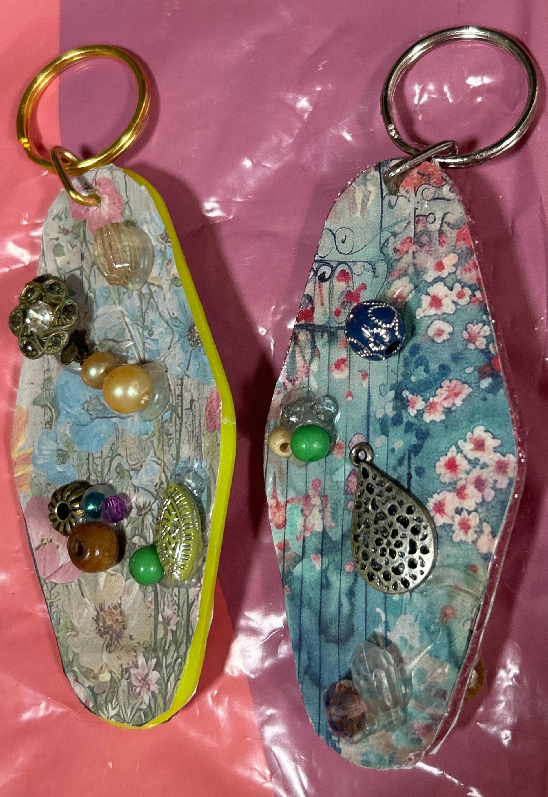 Two decorative keychains on a pink surface with a clear plastic bag in the background.