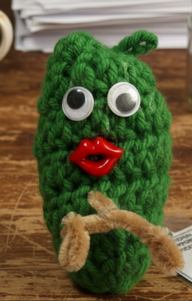 Knitted pickle ornament with googly eyes and a red button on a pink background