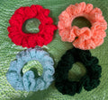 Four crocheted scrunchies in pink, black, red, and blue on a textured green surface.
