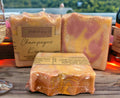 Three wrapped soaps with visible branding on a wooden surface, with a scenic background.