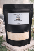 Black pouch of Duckville Soap Co. Mineral Bath Soak on a towel with eucalyptus leaves and candles in the background.