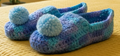 Blue and purple crocheted slippers with pom-poms on a pink blanket.