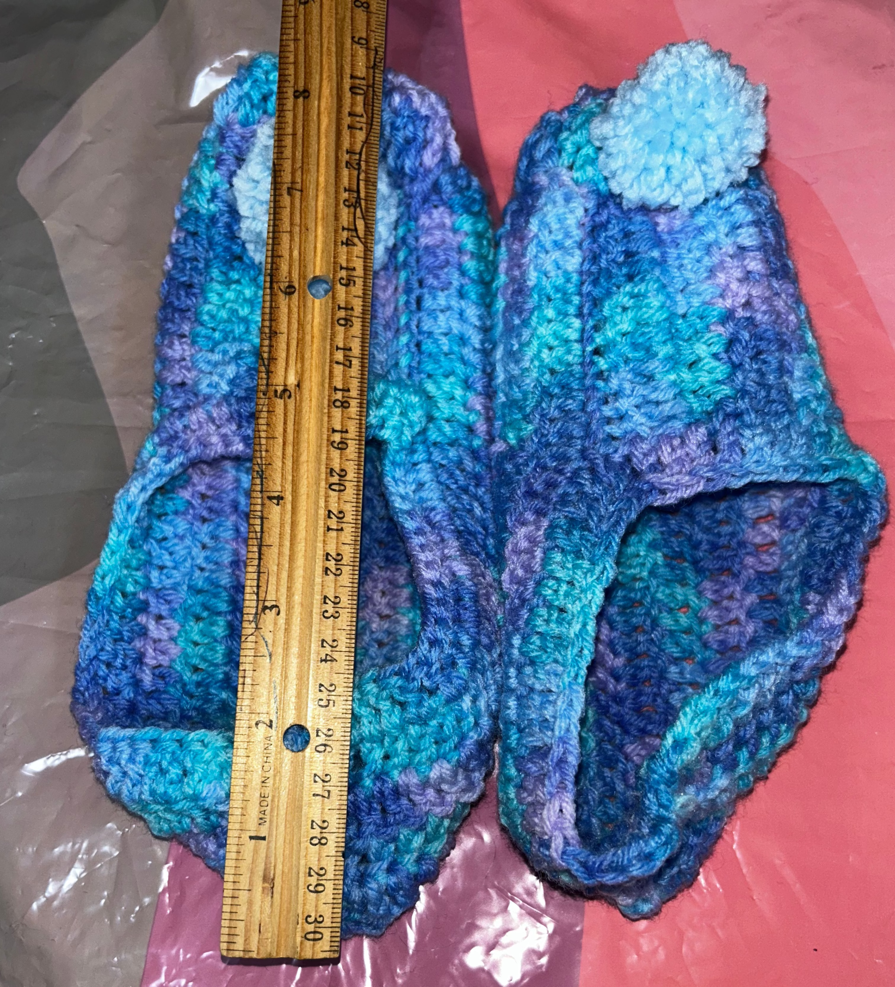 Blue crocheted slippers with a pom-pom on a pink surface, measured by a ruler.