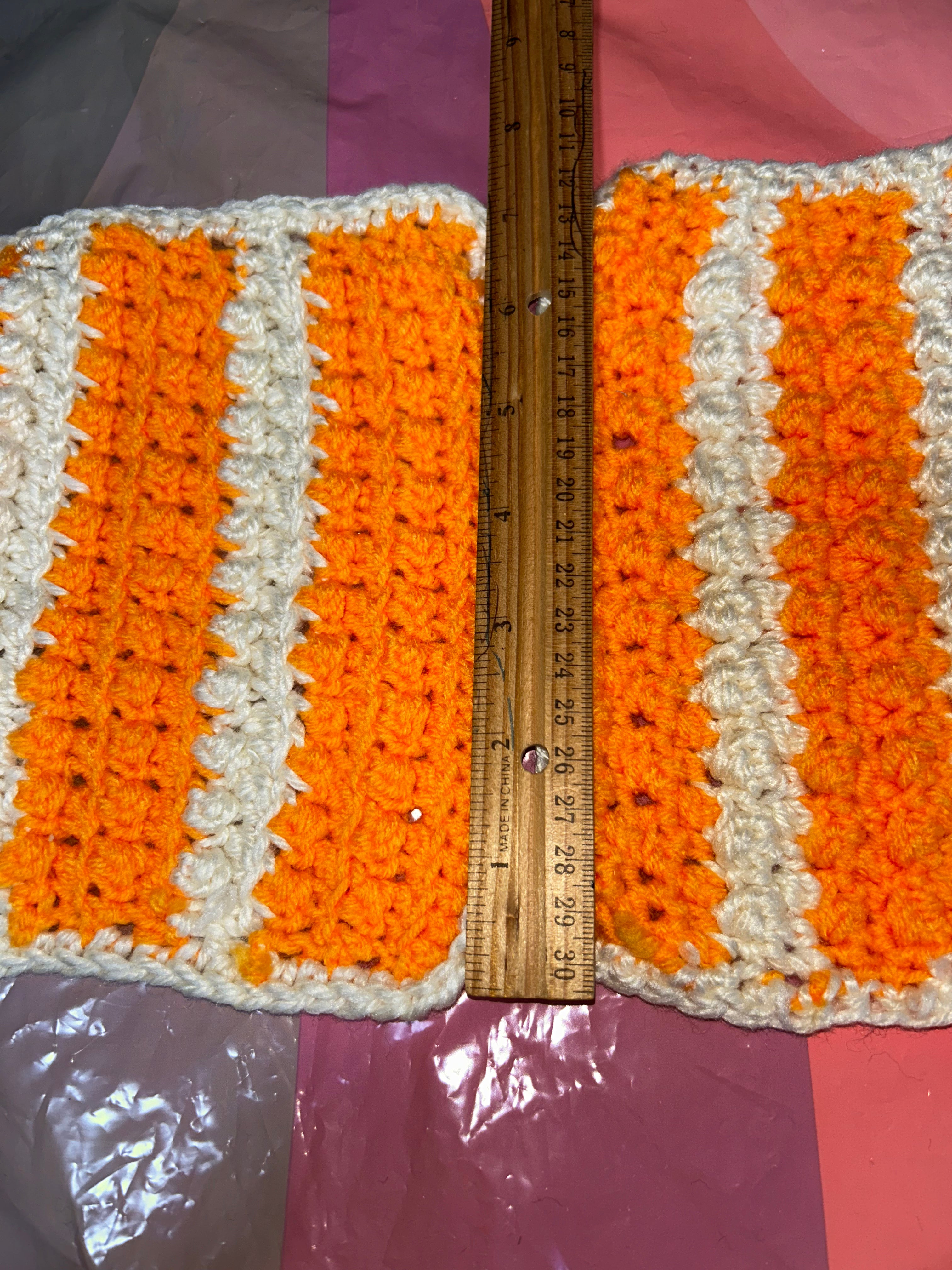 Crocheted potholder with orange and white stripes measured by a ruler on a purple background