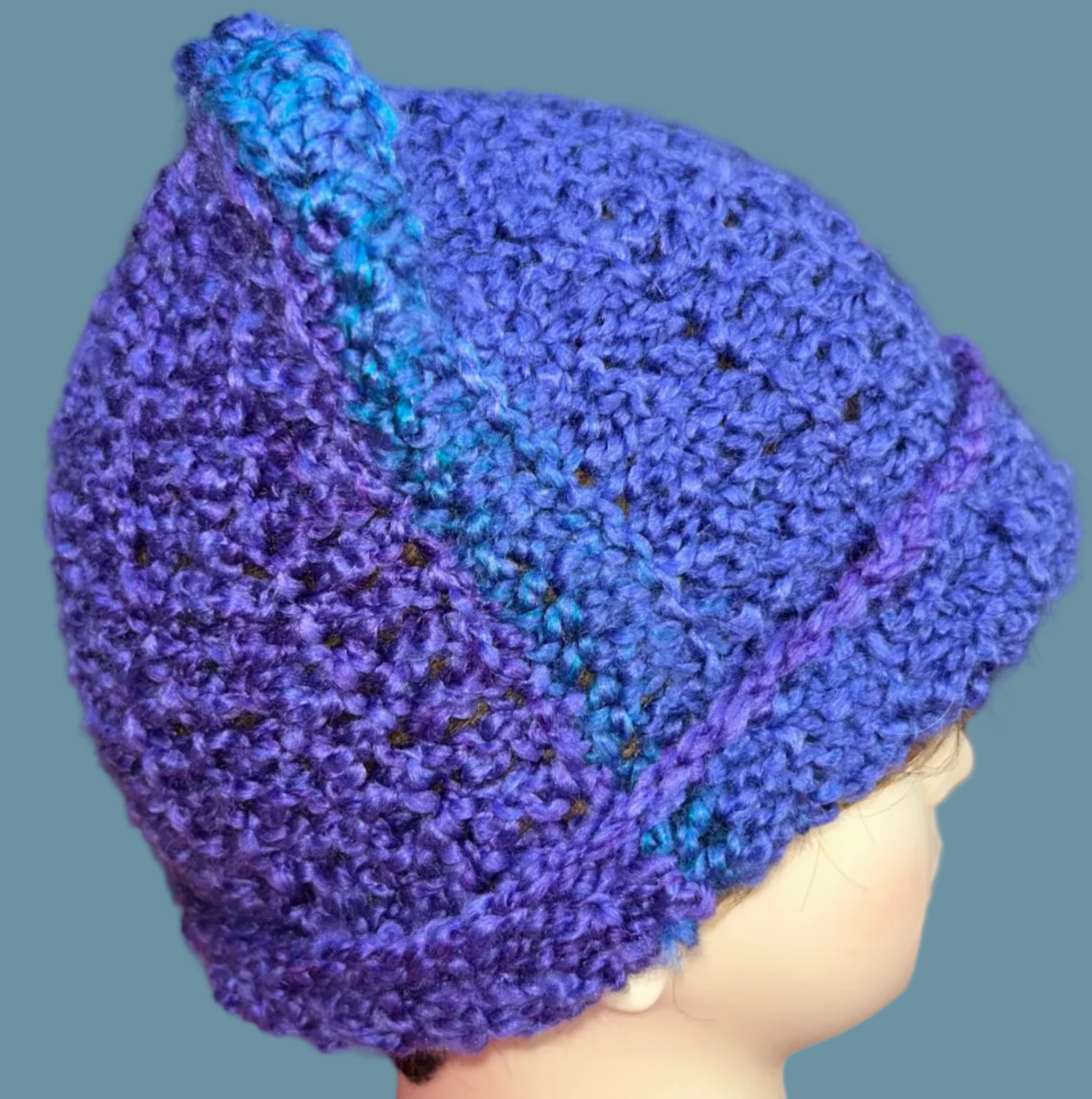 Blue crocheted beanie on a mannequin head with a colorful rug in the background