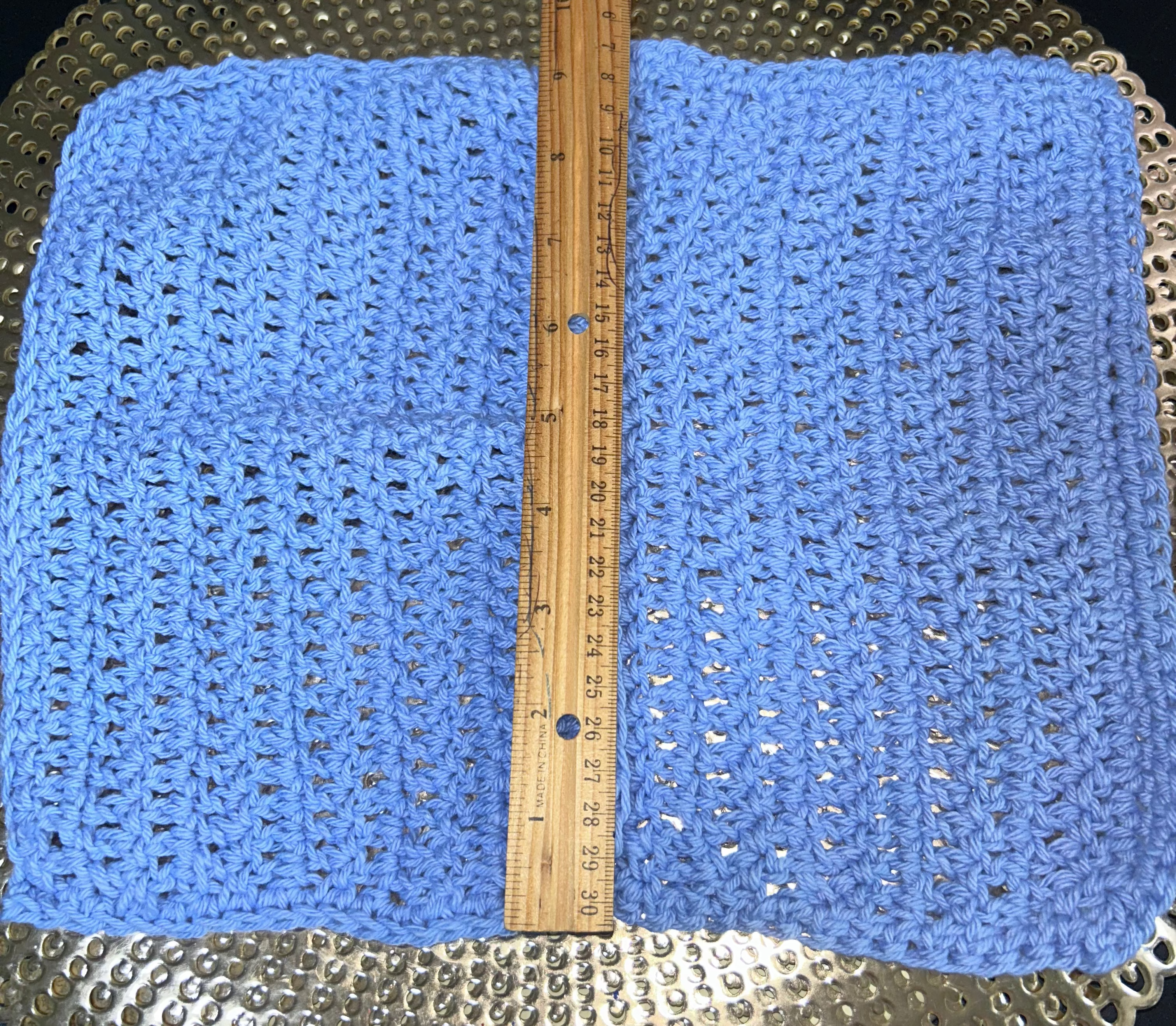 Blue crocheted fabric with a wooden ruler on top, placed on a textured surface.