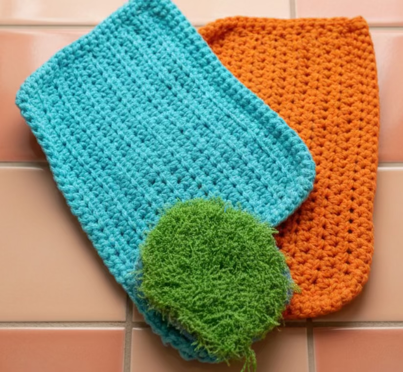Two crocheted dishcloths, one blue and one orange, with a green scrubber on a metallic surface.