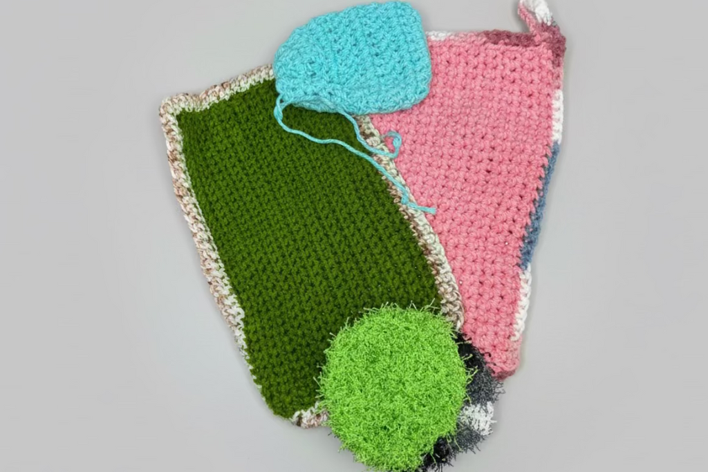 Three knitted items on a textured surface, including a green square, pink triangle, and blue circle.