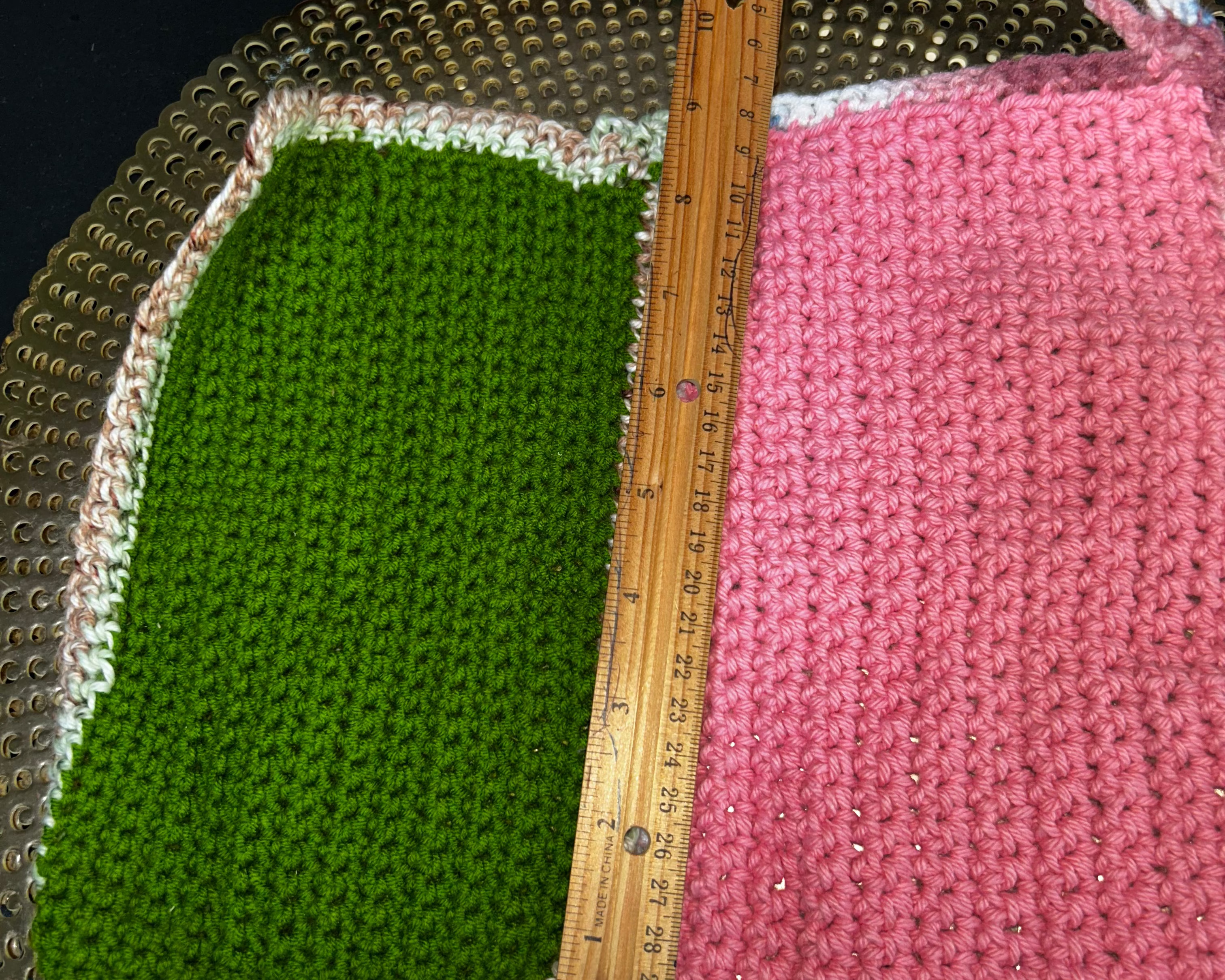 Two crocheted squares, one green and one pink, with a wooden ruler on a metallic surface.