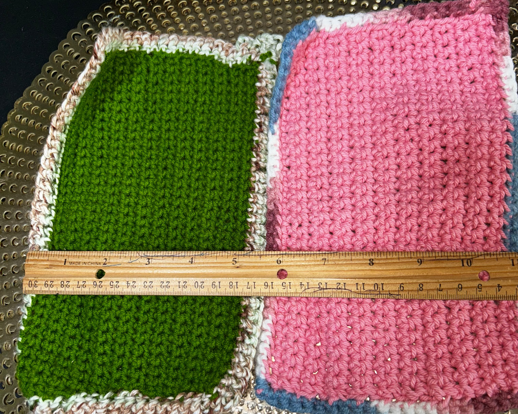Two crocheted squares, one green and one pink with white and blue accents, on a textured surface with a ruler for scale.