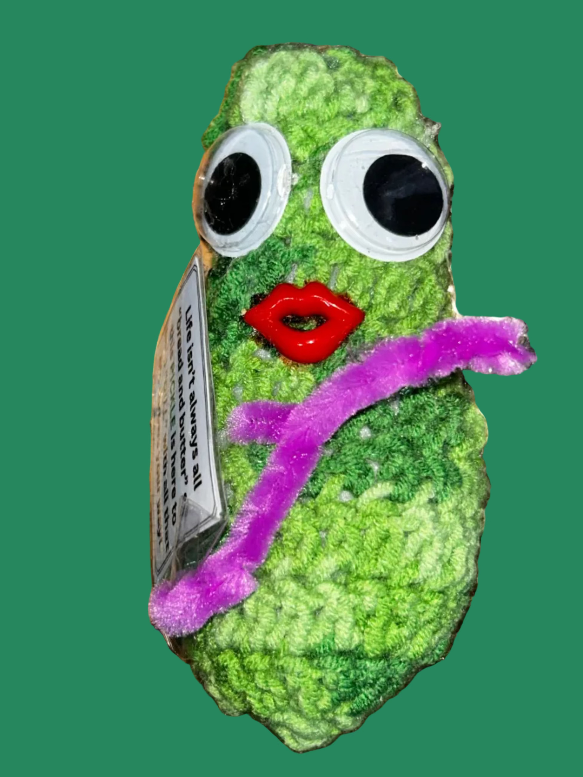 Green knitted toy with googly eyes and a red mouth on a textured metallic surface.