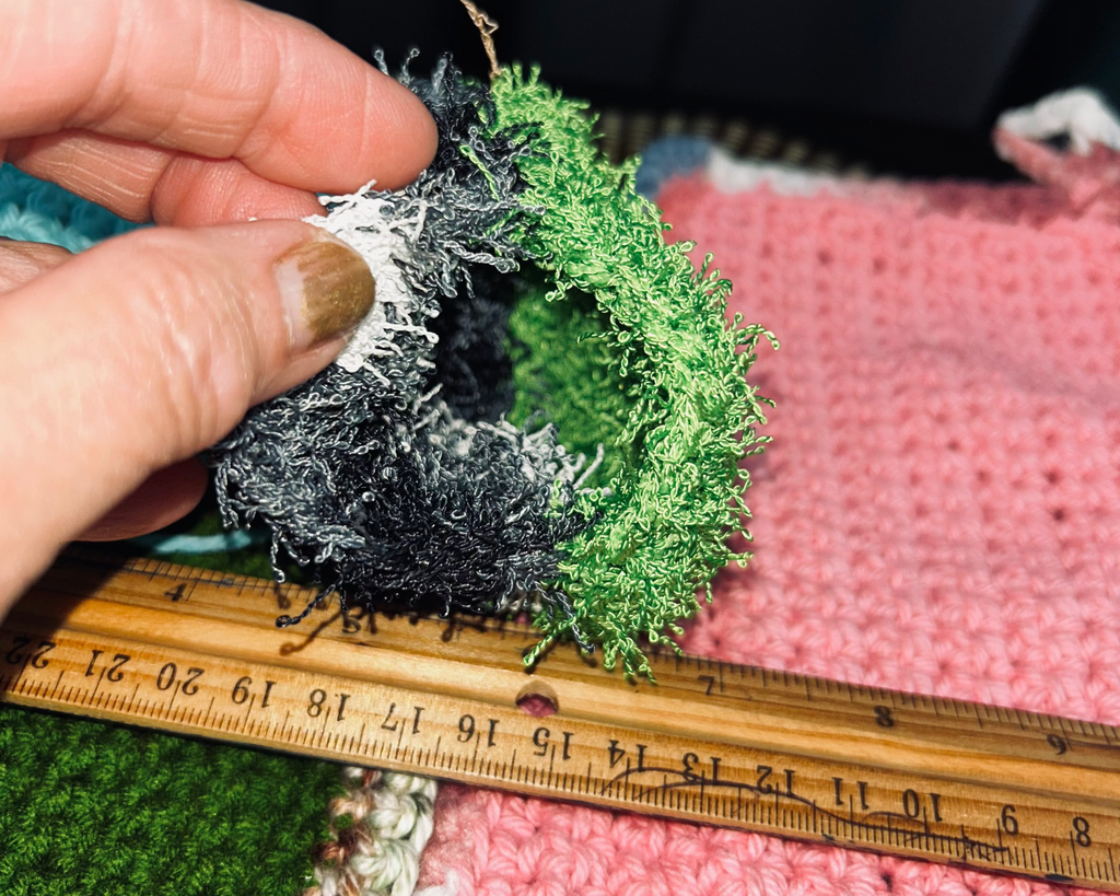 Hand holding green and black textured fabric with a ruler for scale on a pink knitted background