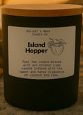 Candle labeled 'Island Hopper' on a sandy surface with shells and a sunset background