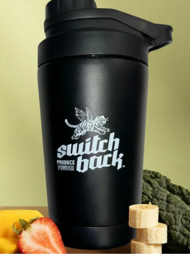 Black shaker bottle with 'switch back' logo on a wooden surface with fruits and vegetables.