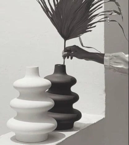 Two vases, one white and one black, on a light surface with a hand holding a plant.