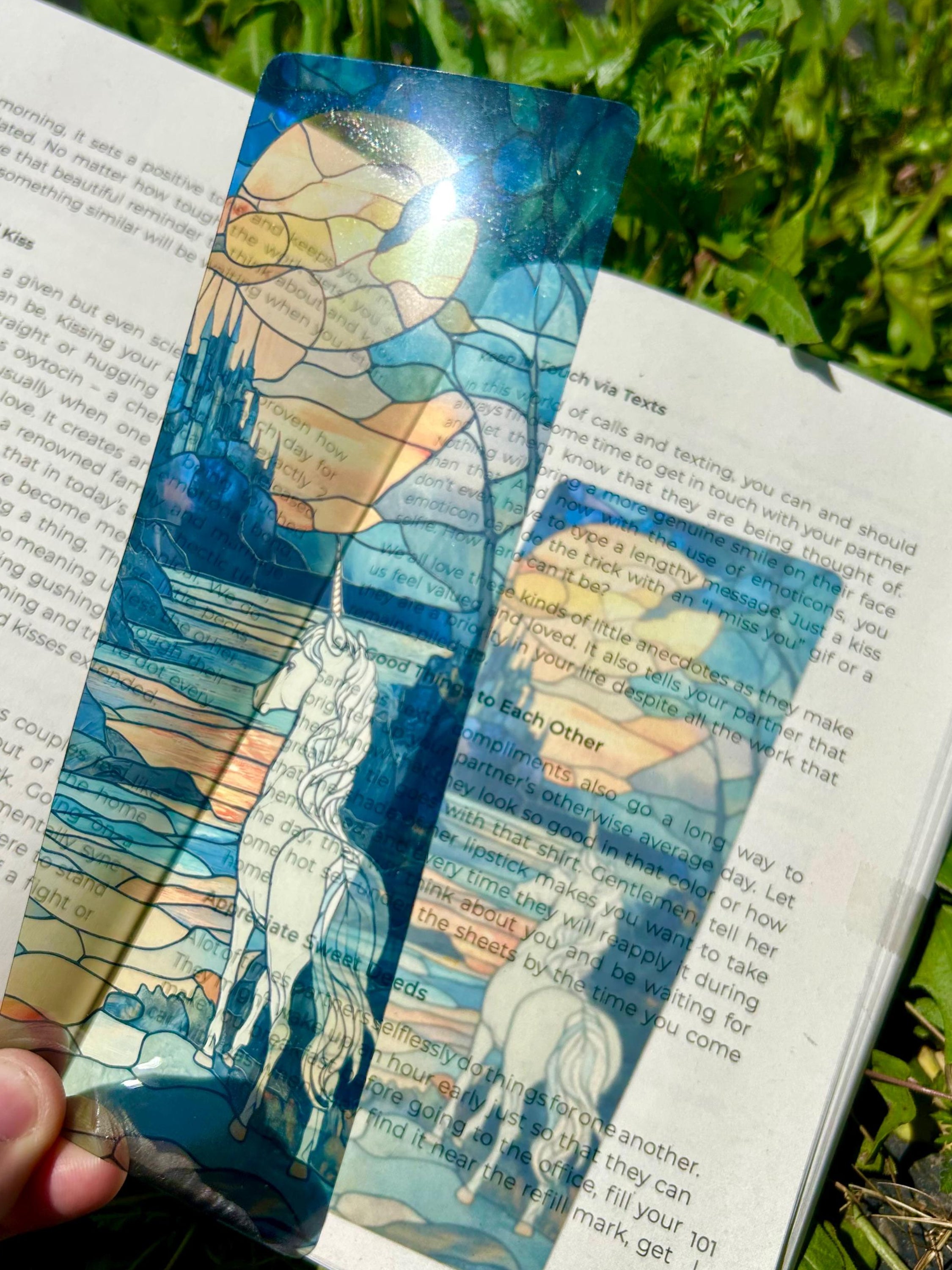 Artistic bookmark with unicorn design held over an open book on green grass